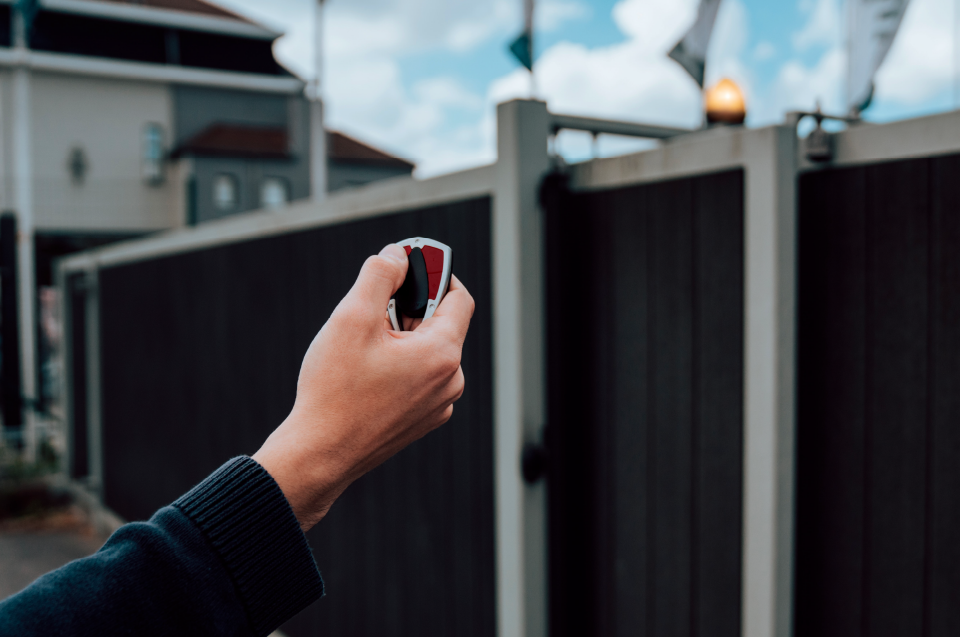 In front of a black automatic gate, a hand is extended holding a small transmitter that when pressed, automatically opens the gates