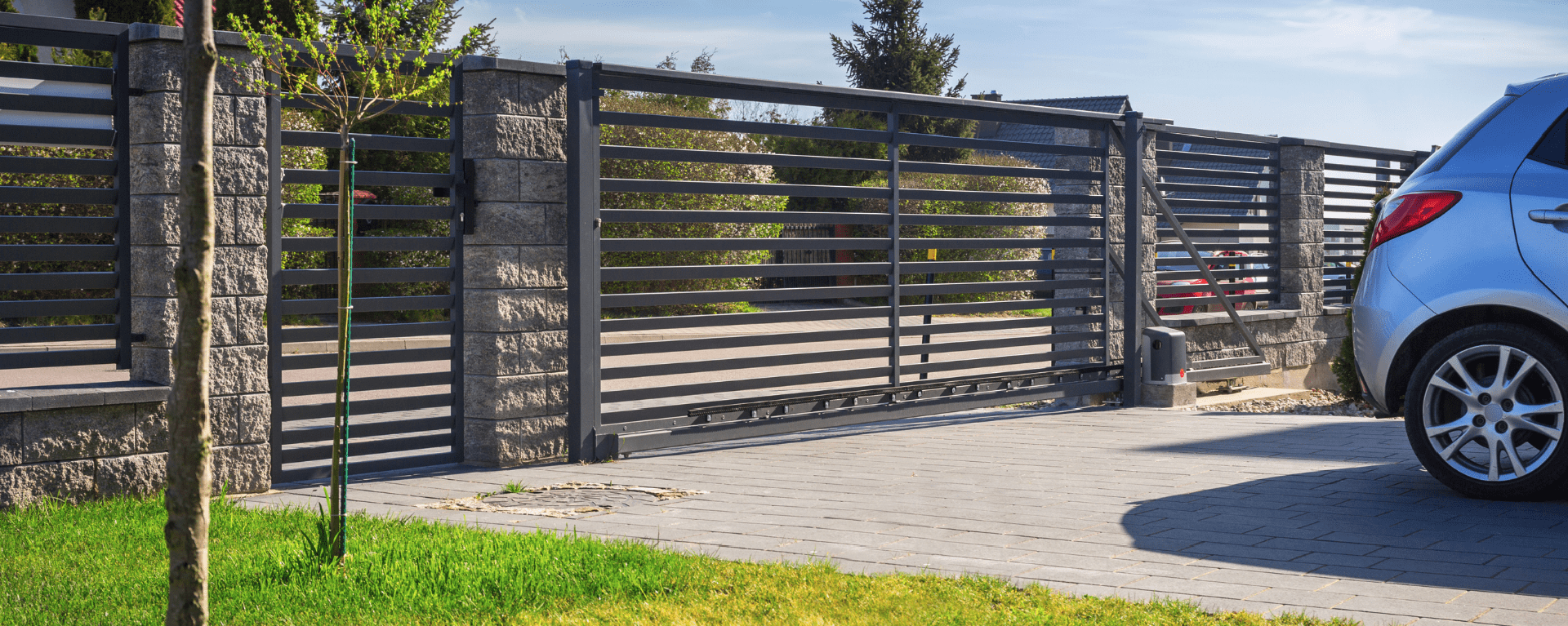 Outside a house on a sunny day, large black automatic gates. The back of a car is seen driving away from the gates.