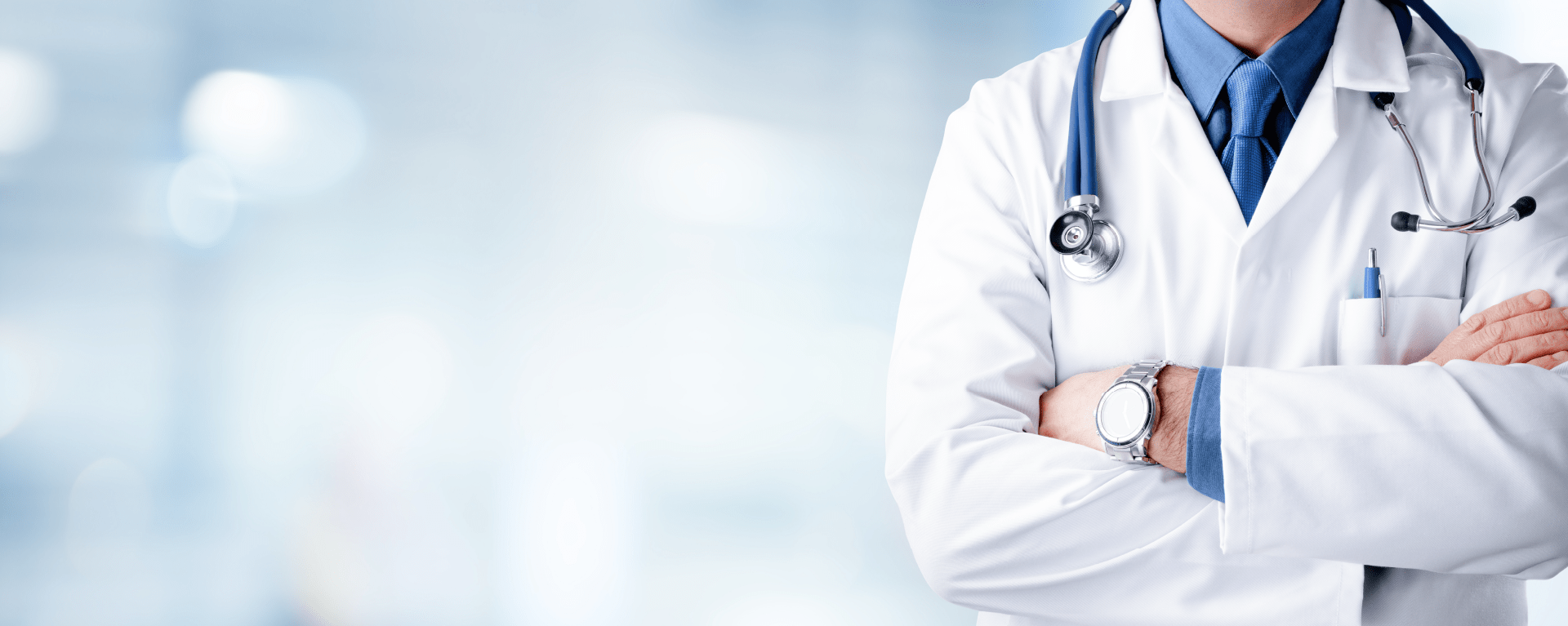 A doctor in a white lab coat with a stethoscope around his neck stands with arms folded in front of a blurred clinical background